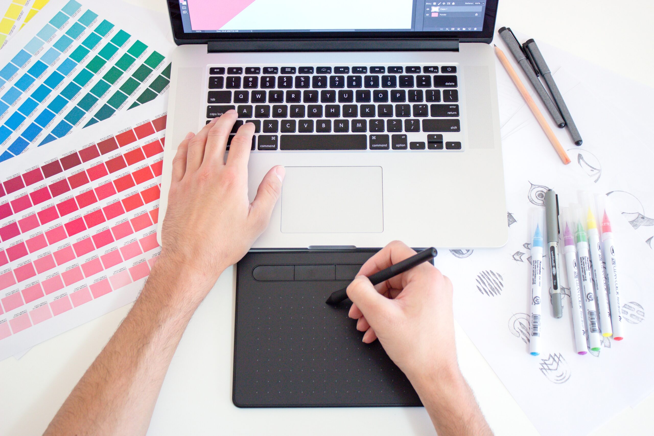 Graphic designer working on a Macbook laptop using a trackpad, color charts and markers